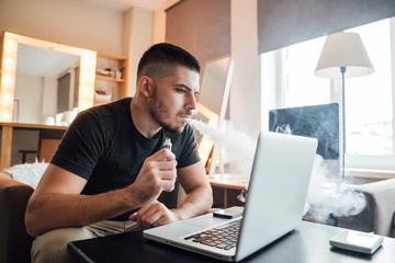 Young businessman working in front of computer in home and smokes an electronic cigarette - freelance and technology concept.