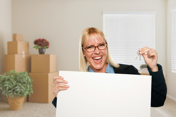 Happy Attractive Woman with Blank Sign and House Key in Empty Room with Packed Moving Boxes and Potted Plants.