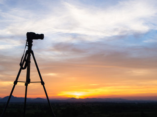 A camera set on a tripod aimed at a silhouette of a landscape at sunset