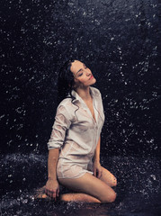 Pretty brunette woman in white shirt under the water drops in aquastudio sitting in water on the knees