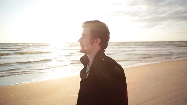 A young dark-haired man in a black coat is walking along the sea shore enjoying the view of the sea.