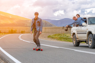 Man rides a longboard in the mountains. Near riding cameraman in SUV car. He takes out the window with an electronic steadycam.