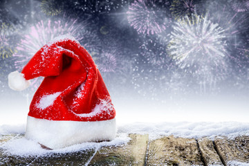 Weihnachtsmütze auf verschneiten Holzbrettern vor Silvesterfeuerwerk