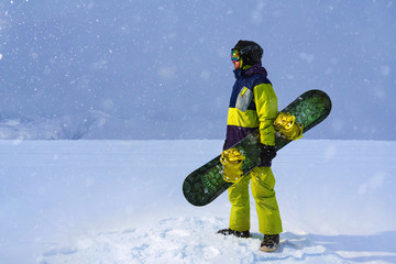 Snowboarder carries a board in  hands. Evening snowfall in  mountains.