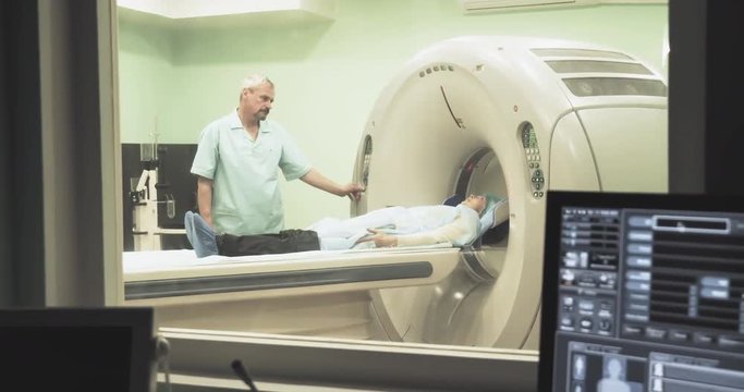 Female patient on CT MRI scanner with doctor 4k video. Computer screen with result diagnosis image. View through glass window in hospital
