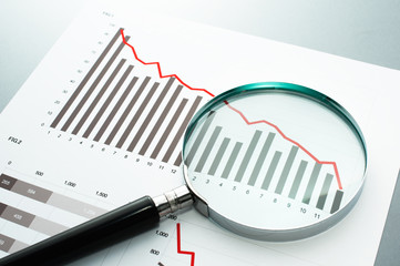Reviewing financial report. Looking at decrease chart with magnifying glass.
Document, chart and magnifying glass on gray reflection background.

