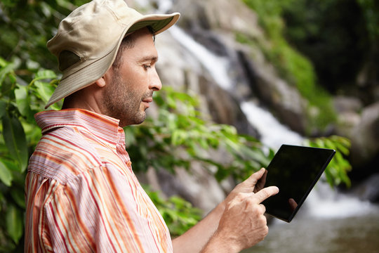 Profile of serious male scientist with stubble taking picture of nature on his black generic digital tablet while working on scientific research in jungle. Botanist using electronic gadget outdoors