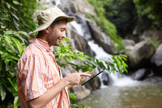 Bearded botanist wearing panama hat using digital tablet pointing at blank screen with happy expression, standing at mountain river against waterfall background while conducting water analysis