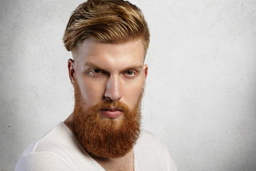 Close up portrait of fashionable redhead hipster man with fuzzy beard and trendy haircut wearing white t-shirt while posing isolated against studio wall background with serious or angry expression