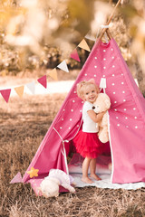 Smiling baby girl 2-3 year old holding toy teddy bear playing in wigwam outdoors. Looking at camera. Childhood. © morrowlight