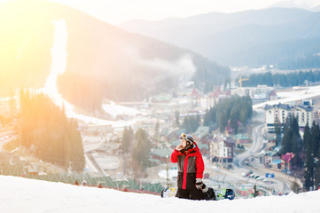 Male snowboarder sitting on ski slope, he's kneeling, looking away and talking on the phone, with an astonishing view on mountains and a small town located between the hills