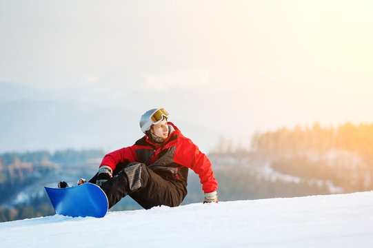 Snowboarder wearing helmet, red jacket, gloves and pants sitting on snowy slope on top of a mountain looking away, with an astonishing view on hills. Carpathian mountains