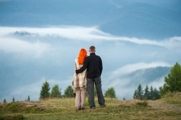 Fototapeta na wymiar Romantic couple standing on a hill enjoying a morning haze over the mountains. Man hugging red-haired woman, she is covered with a plaid. Back view