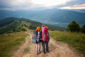 Obraz premium Young couple hikers with backpacks on the road in the mountains. On the background - mountains, forests, hills, village in the valley and cloudy sky