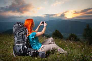 Happy tourist woman with a backpack sitting in the grass with wild flowers on a hill and makes the photo of mountains on the phone. Incredible view of beautiful mountains and cloudy sky at sunset