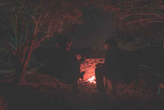 Couple sitting at burning camp fire in the night. Camping in the forest under starry sky, Namibia, Africa. Summer adventures and exploration in the african National Parks. Toned image.