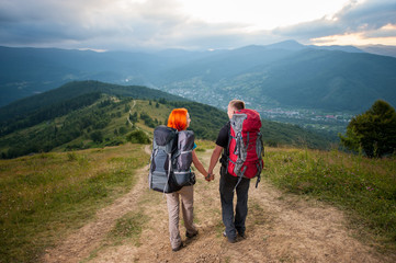 Fototapeta na wymiar Back view of happy man and red-haired woman walking, holding hands, smiling. Couple with backpacks on the road in the mountains. Forests, hills, village in the valley and cloudy sky on the background