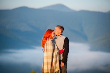 Naklejka premium Romantic couple tourists kissing and hugging each other against beautiful mountain landscape with morning haze over the mountains on background. Red-haired girl covered with a blanket. Close-up