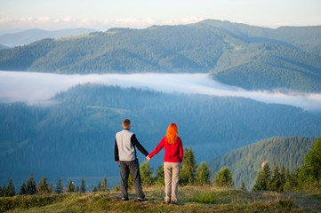 Rear view man holding hands red-haired girl standing on a hill, enjoying a morning haze over the beautiful mighty mountains