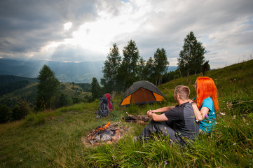Loving couple hikers sitting on the green grass near campfire, tent, backpacks and looking into the distance on the hills, mountains, forest and punching the sun's rays through a cloudy sky at sunset