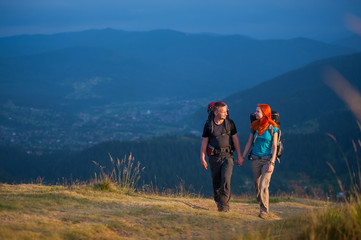 Obraz premium Travelers man and woman with backpacks climbing up in the beautiful mountains area, holding hands and looking to each other. Lifestyle active vacations concept mountains landscape on background