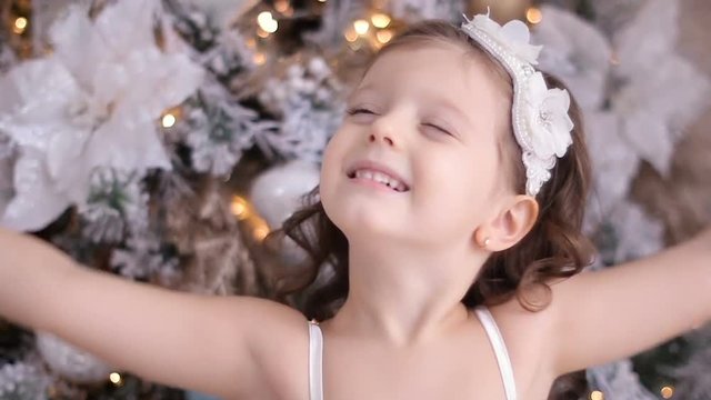 little girl three years old in a white dress smiling at the decorated Christmas tree in room
