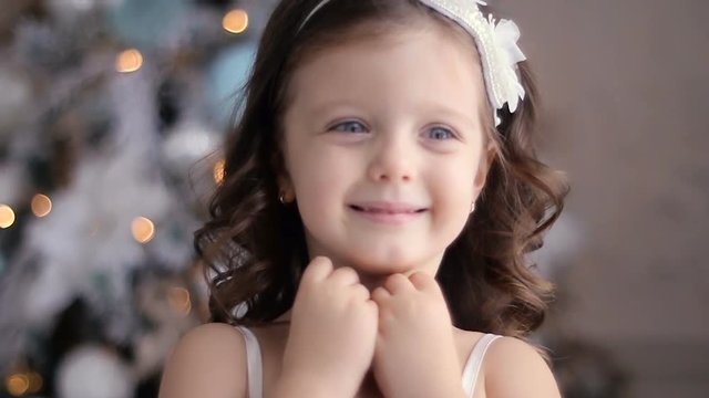 a little girl three years old in a white dress smiles and sends a kiss by the decorated Christmas tree in room