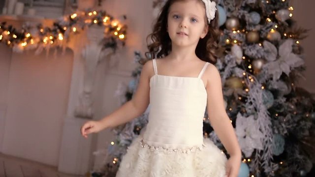 little girl three years old in a white dress dancing around the decorated Christmas tree in room