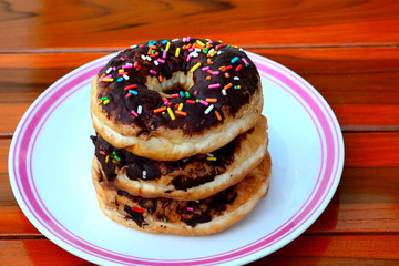 Overhead view of a plate of iced chocolate sprinkle ring donuts on a rustic wooden table background.