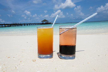 Two colorful cocktails laying on white sand, small corals covered beach in front of the amazing turquoise water of the indian ocean.