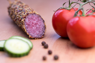 Delicious smoked sausage whole on wooden board with slices of cucumbers, tomatoes. View from above.