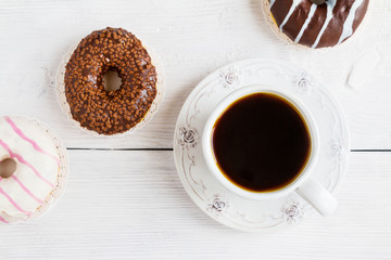Chocolate and glazed donuts, pink, white, brown  on white wooden table and cup of black coffee, top view.