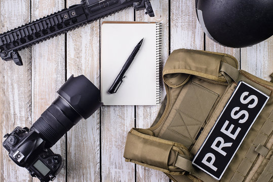 Body armor for photojournalist,digital camera,notebook and rifle/Bulletproof vest with patch "PRESS",notebook,DSLR camera,helmet and rifle on wooden table.Top view.