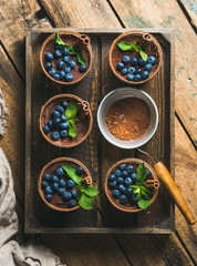 Homemade Tiramisu dessert in glasses with cinnamon sticks, mint leaves, fresh blueberries and sieve with cocoa powder in wooden tray over rustic wooden background, top view, vertical composition