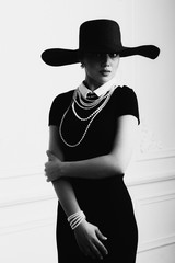Elegant beautiful young woman in retro style dress, hat standing over light grey background. Black and white monochrome photo