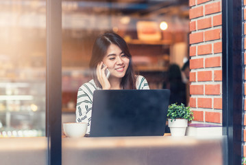 Viewed through glass with reflections of happy beautiful young Asian woman smile while talking on the mobile phone with laptop at cafe