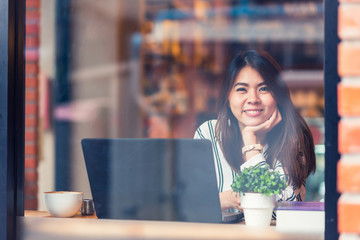 Viewed through glass with reflections of happy beautiful young Asian woman smile while working with laptop at cafe
