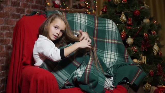 Girl straightens her hair sitting in a chair near a Christmas tree