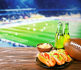 Beer with snack and ball on wooden table against football field background. Sport and entertainment concept.