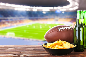 Beer with snack and ball on wooden table against football field background. Sport and entertainment concept.