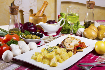 Fried fish steak with side dish on a white plate beside fresh vegetables, mushrooms, lemon, olive oil, olives, sauce and white wine.