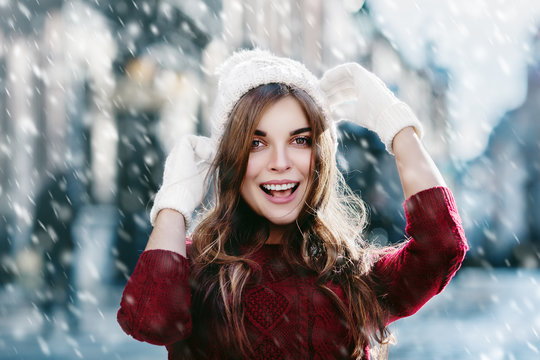  Outdoor close up photo of beautiful girl acting thrilled, wearing stylish knitted winter hat and gloves. Model expressing joy and looking at camera. Christmas, New Year concept. Magic snowfall effect