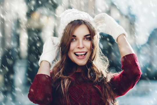 Outdoor close up portrait of young beautiful happy smiling girl, wearing stylish knitted winter hat and gloves. Model and looking at camera. Christmas, New Year concept. Magic snowfall effect. Toned