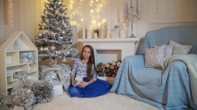 Girl sitting under the tree by the fireplace and shaking a box with a gift