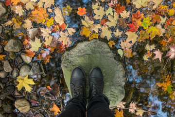 Autumn leaves floating in the stream past woman's feet. Looking down at feet in black boots standing on a rock in the middle of a stream full of fall leaves