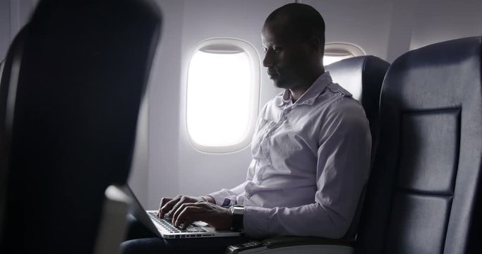 African American man using laptop computer and looking out of window in main cabin of commercial airliner.  Medium long shot from side angle, recorded hand held at 60fps.