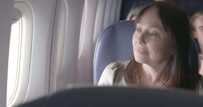 View across seats of mature woman sitting next to window and looking out from main cabin of commercial airliner. Close up from front, recorded hand held at 60fps.