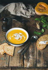 Pumpkin cream soup in white bowl with fresh basil, spices and grilled bread slices over old rustic wooden background, copy space, selective focus, vertical composition