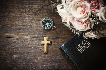 the wooden cross with compass and holy bible and roses flowers on wooden background, vintage tone...