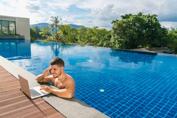 Workplace at the resort. Young man with a phone and a laptop in the swimming pool. Freelancer at...